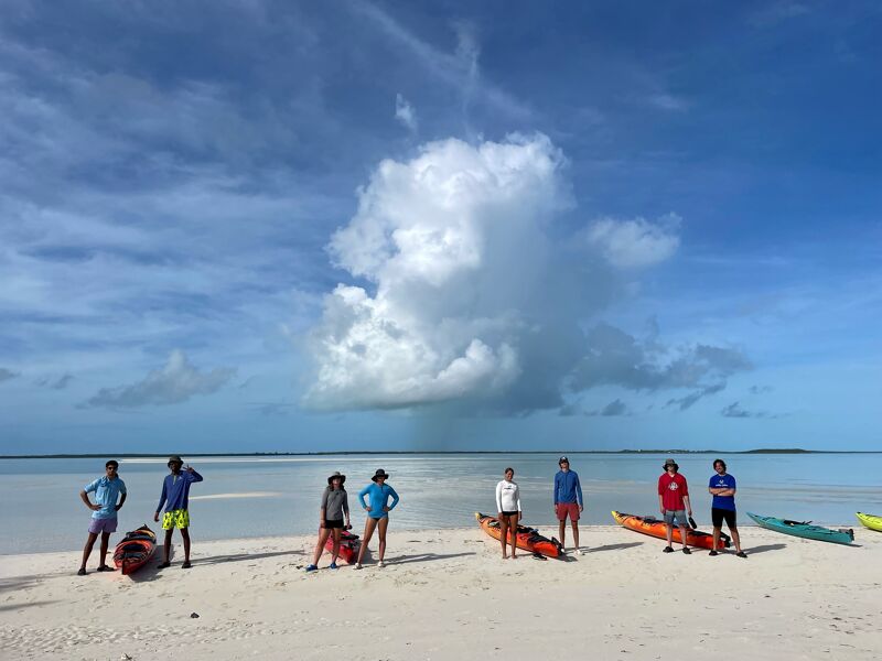 The image shows a group of people standing on a sandy beach with kayaks. The sky is blue with some clouds. The water is calm and clear. The people are dressed in casual clothes and appear to be enjoying a day at the beach.